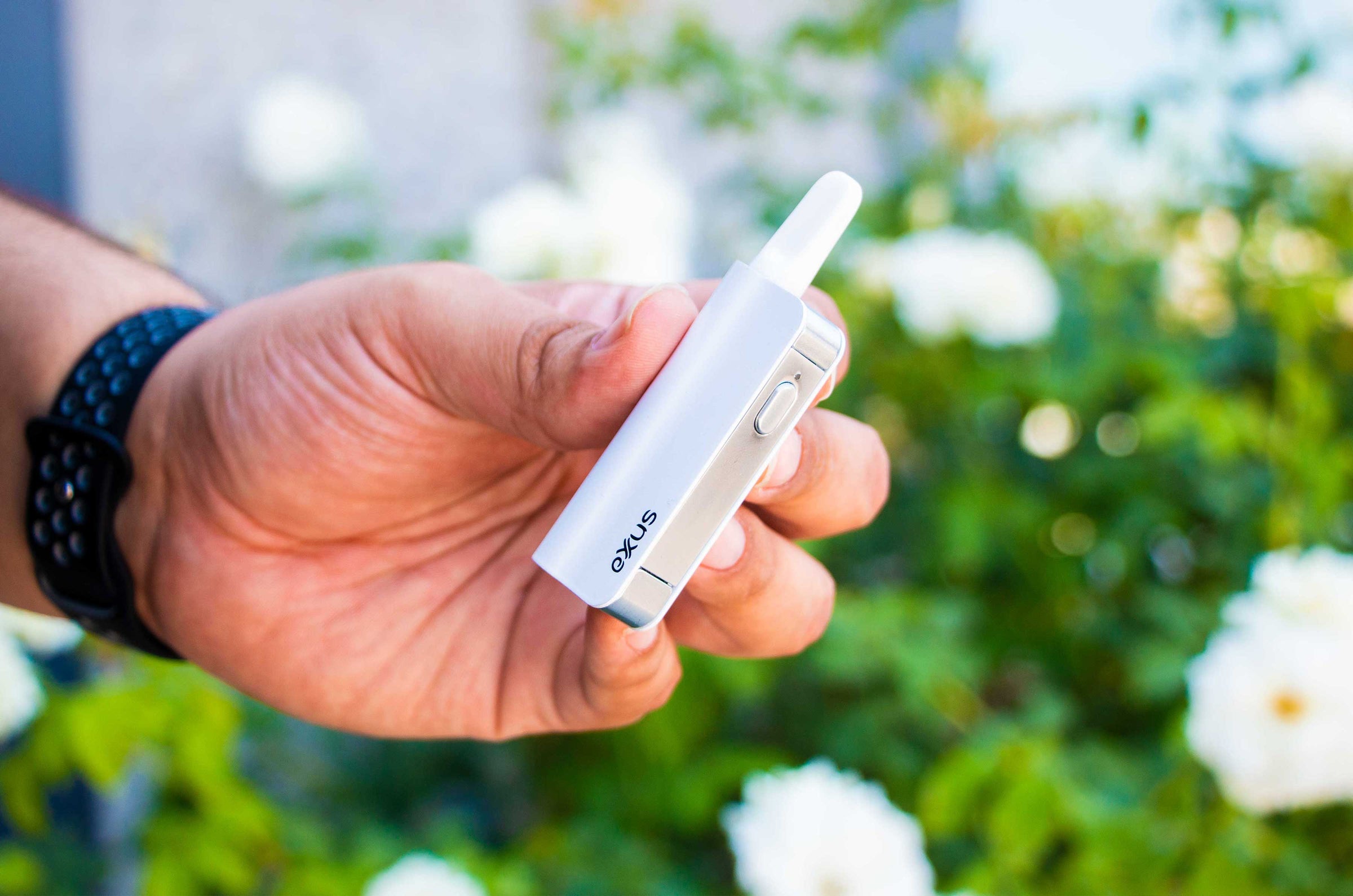 man holding a white Exxus Snap VV Mini portable Vaporizer outside near flower bush