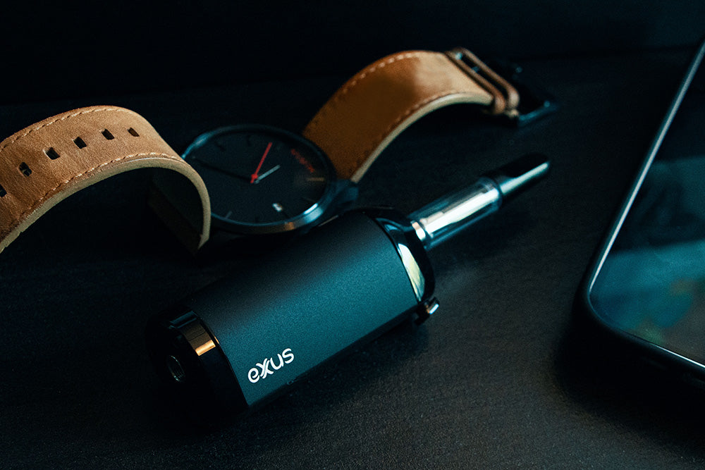 Black Exxus VRS concentrate vaporizer on a desk beside a watch