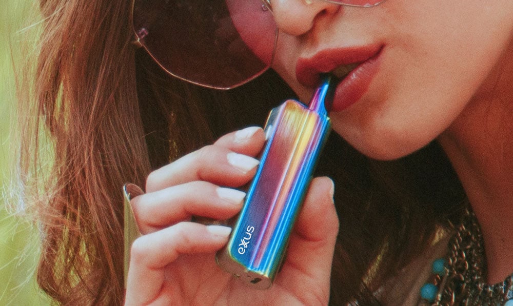 Woman vaping Exxus Snap VV in front of forest under natural lighting