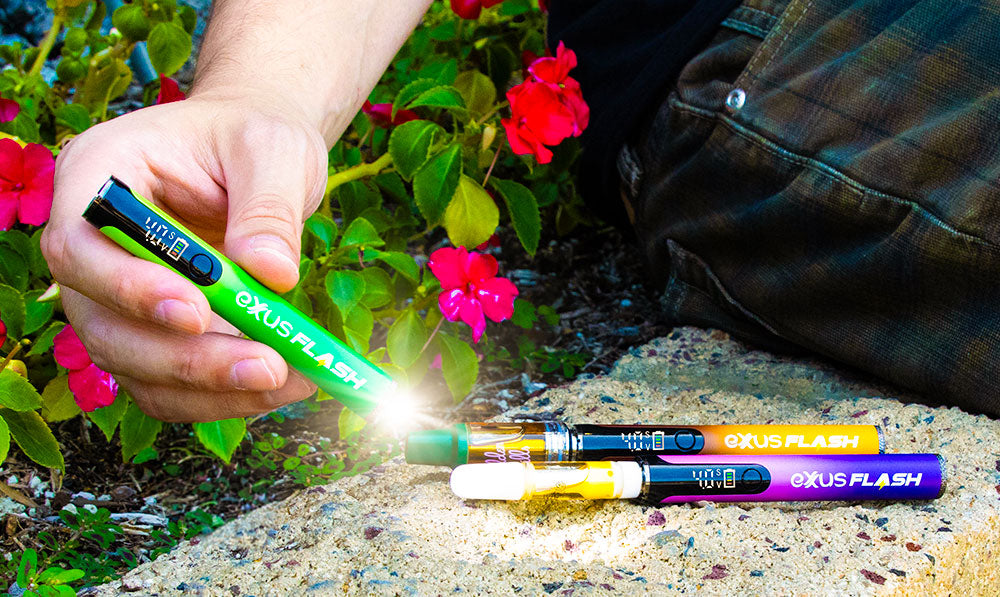Four Exxus Flash cartridge vaporizers in green, orange, yellow and blue on a stone ledge beside pink flowers